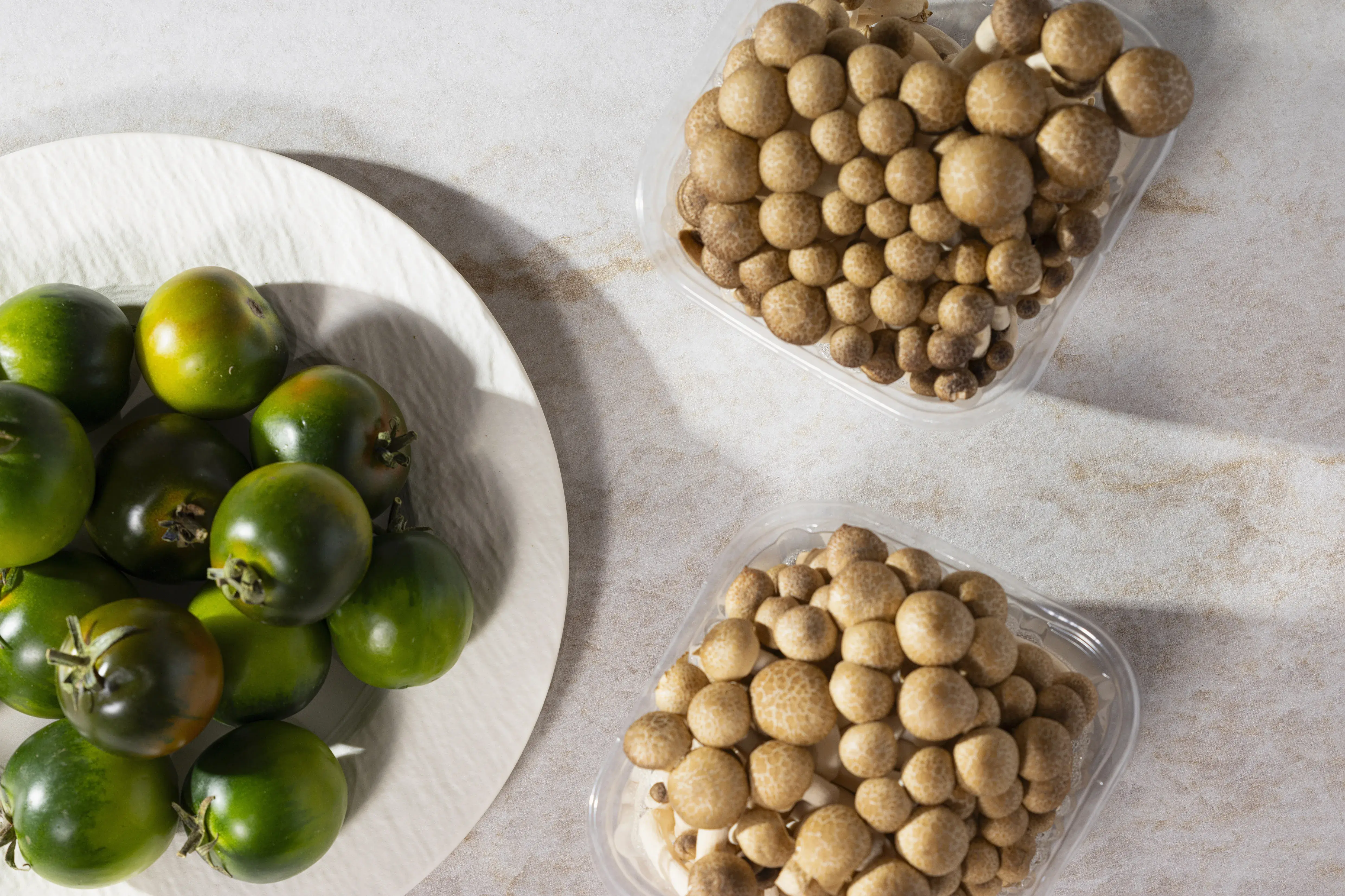 Grüne Tomaten auf einem Teller, daneben zwei Schalen mit braunen Champignons.
